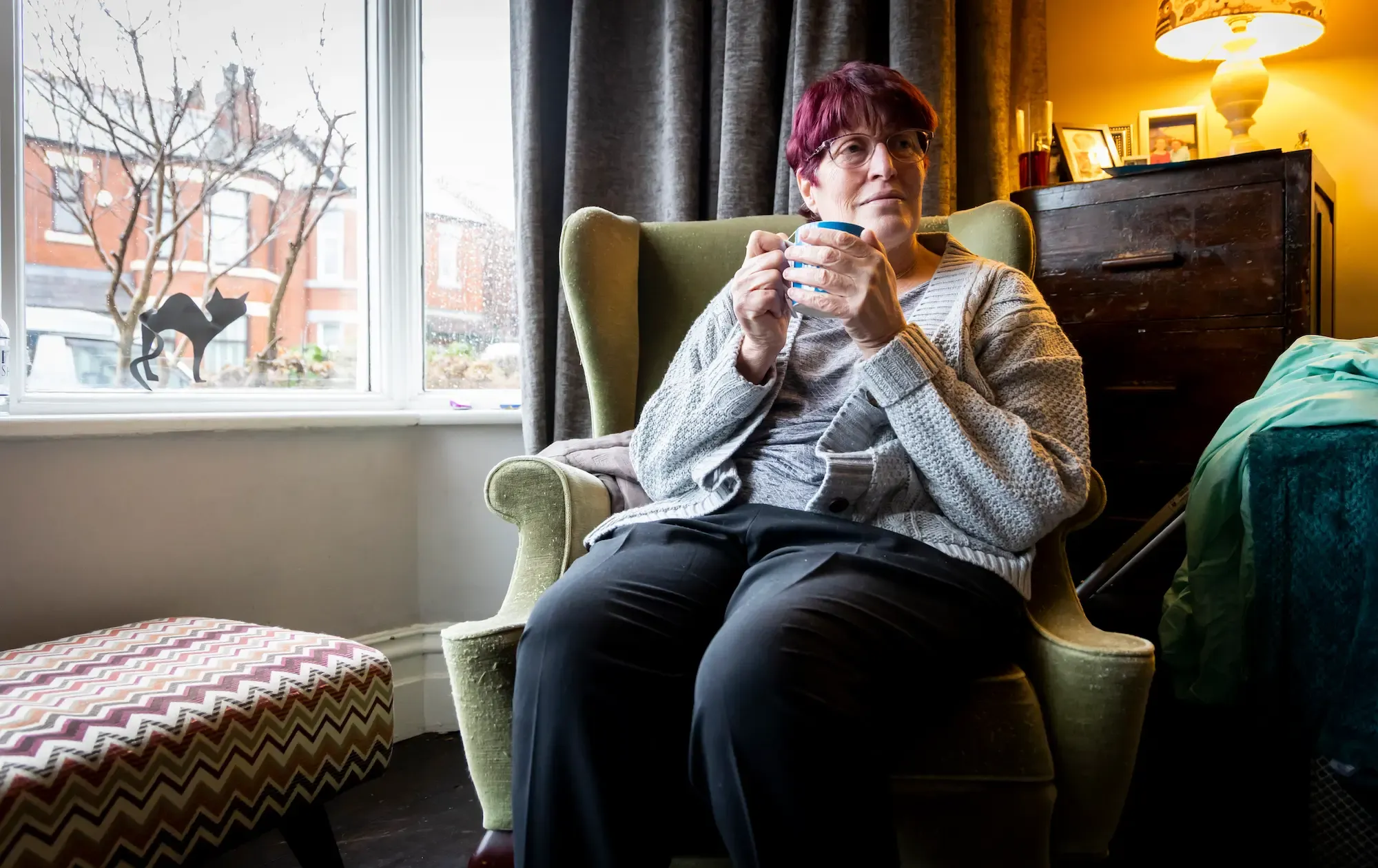 Middle-aged woman sat by a window drinking from a mug